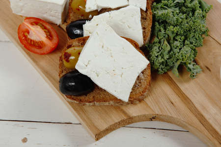 fresh greek feta cheese sandwich on wooden plate with hot green olives and black, goat cube, tomatoes kale over tableの写真素材