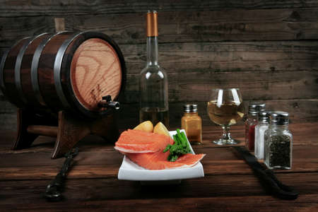 fresh raw salmon fillet served with lemon and white wine in wineglass, wooden barrel, bottle, on white plate over vintage wood table with handmade cutlery knife and fork with empty space backgroundの写真素材