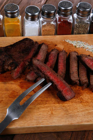 grilled beef fillet steak on fork over wooden board served with different kind of dry spices salt crushed black pepper hot baked sweet paprika red hot chili on dark tableの写真素材