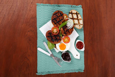 junk food meat big beef hamburger fried eggs on white  wood plate with cutlery ketchup sauce and pickels on blue mat over wooden tableの写真素材
