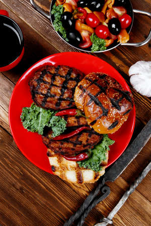 fresh grilled beef hamburger served on red plate with black coffee glass chili pepper olives green kale leaves mushroom bell garlic and forged vintage antique cutlery over wooden tableの写真素材