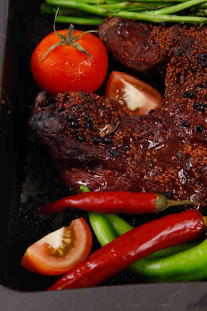 fresh glazed baked big beef meat rib chunk under sweet sauce with tomatoes hot chili pepper asparagus different peppercorn spices in black deep tray on light walnut wooden tableの写真素材