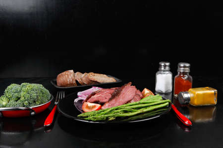 business lunch fresh roast beef meat slices on black plate wotj cutlery asparagus boiled broccoli rye bun on wooden tableの写真素材
