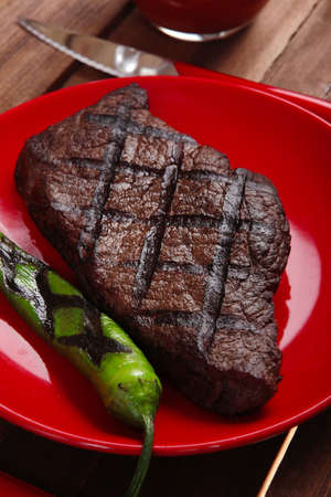 red theme lunch : fresh grilled bbq roast beef steak on red plate with green chili tomato soup ketchup sauce paprika small jug glass ground pepper american peppercorn and modern cutlery served on wooden tableの写真素材