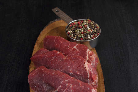 fresh raw beef piece fillet on middle size wooden plate with pepper and peppercorns in measuring spoon space for text on black backgroundの写真素材