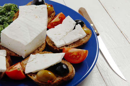 white feta greek cheese sandwich rye bread on blue plate with black spain olives tomatoes and cutlery over retro wooden tableの写真素材