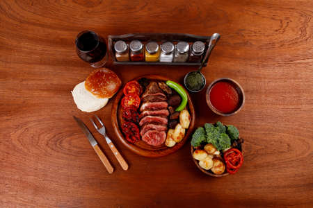 fresh roast bbq beef meat ribeye steak on wooden plate served with tomato juice in wooden cup, boiled broccoli, baked tomatoes and potatoes, with white bun, and red wine glass on light walnut wooden tableの写真素材