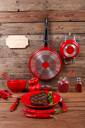 red theme lunch : fresh grilled bbq roast beef steak on red plate with green chili tomato soup ketchup sauce paprika small jug glass ground pepper american peppercorn and modern cutlery served on wooden table with empty nameplateの写真素材