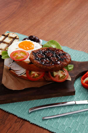 junk food big beef hamburger fried eggs on dark wood plate with modern cutlery ketchup sauce and pickels on blue mat isolated over white backgroundの写真素材