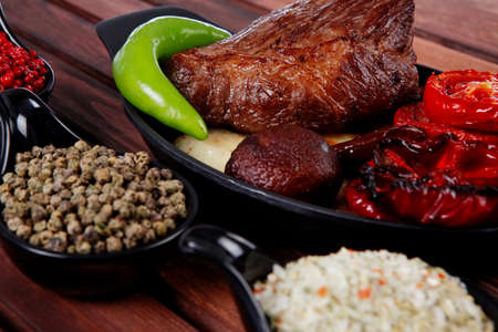 fresh roast bbq beef steak on black pan baked with mushroom potatoes tomatoes green hot chili pepper served with different peppercorn on dark wooden tableの写真素材