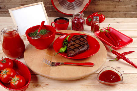 red theme lunch : fresh grilled bbq roast beef steak on red plate with green chili tomato soup ketchup sauce paprika small jug glass ground pepper american peppercorn and modern cutlery served on wooden plate over table empty nameplate menu boardの写真素材