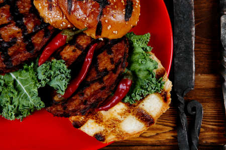 fresh grilled beef hamburger served on red plate with black coffee glass chili pepper olives green kale leaves mushroom bell garlic and forged vintage antique cutlery over wooden tableの写真素材