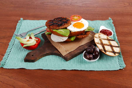 junk food big beef hamburger fried eggs on dark wood plate with modern cutlery ketchup sauce and pickels on blue mat isolated over white backgroundの写真素材