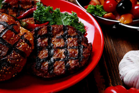 fresh grilled beef hamburger served on red plate with black coffee glass chili pepper olives green kale leaves mushroom bell garlic and forged vintage antique cutlery over wooden tableの写真素材
