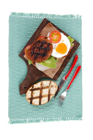 junk food big beef hamburger on dark wood plate with modern cutlery on blue mat isolated over white backgroundの写真素材