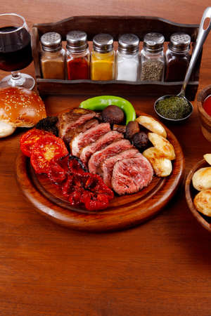 fresh roast bbq beef meat ribeye steak on wooden plate served with tomato juice in wooden cup, boiled broccoli, baked tomatoes and potatoes, with white bun, and red wine glass on light walnut wooden tableの写真素材