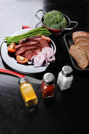 business lunch fresh roast beef meat slices on black plate wotj cutlery asparagus boiled broccoli rye bun on wooden tableの写真素材