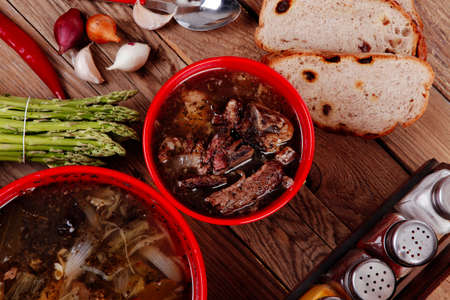 home style beef meat soup vintage country wood table asparagus pepper bell spices bread garlic cutlery spices red bowlsの写真素材