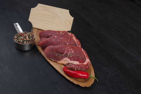fresh raw beef piece fillet on middle size wooden plate with pepper and peppercorns in measuring spoon with name plate space for text on black backgroundの写真素材