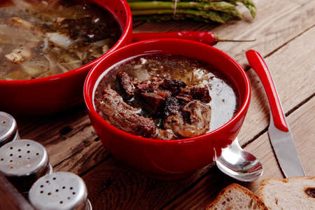 home style beef meat soup vintage country wood table asparagus pepper bell spices bread garlic cutlery spices red bowlsの写真素材