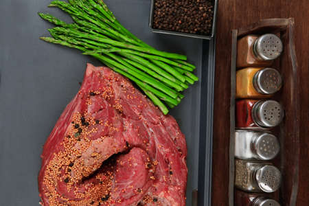meat raw beef fillet chunk on black tray asparagus on wooden table allspice pink white black green peppercorn stainless cutlery knife forkの写真素材