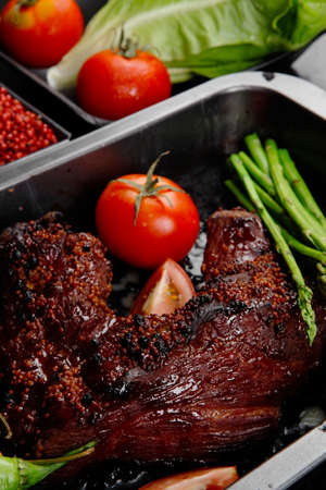 fresh glazed baked big beef meat rib chunk under sweet sauce with lettuce broccoli tomatoes hot chili pepper pink peppercorn different spices on black wooden tableの写真素材