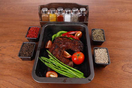 fresh glazed baked big beef meat rib chunk under sweet sauce with tomatoes hot chili pepper asparagus different peppercorn spices in black deep tray on light walnut wooden tableの写真素材