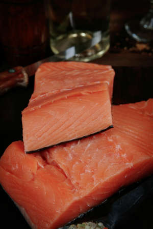 fresh raw salmon fillet served with lemon and white wine in wineglass, bottle, on dark plate over vintage wood table with handmade cutlery knife and spice on spoonの写真素材