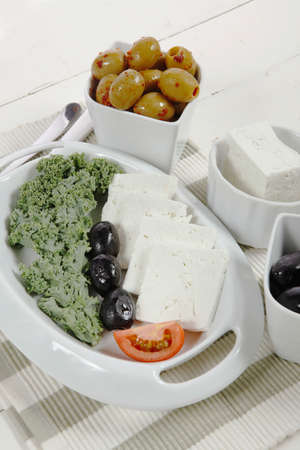 fresh greek feta cheese sandwich on white plate with hot green olives and black, goat cheese cube, tomatoes kale over retro tableの写真素材