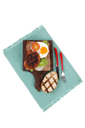 junk food big beef hamburger on dark wood plate with modern cutlery on blue mat isolated over white backgroundの写真素材
