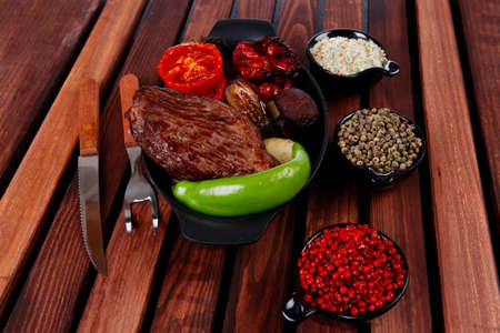 fresh roast bbq beef steak on black pan baked with mushroom potatoes tomatoes green hot chili pepper served with different peppercorn on dark wooden tableの写真素材