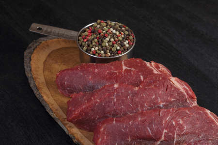 fresh raw beef piece fillet on middle size wooden plate with pepper and peppercorns in measuring spoon space for text on black backgroundの写真素材