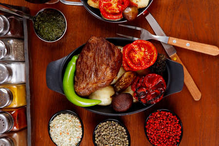 fresh roast beef meat rib eye steak with tomatoes,potatoes, hot chili pepper gern and pink peppercorn saucer, cutlery, set of spice, and red wine glass on light walnut tableの写真素材