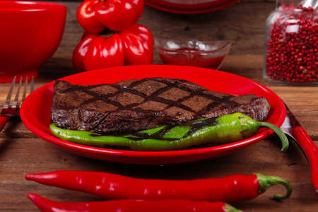 red theme lunch : fresh grilled bbq roast beef steak on red plate with green chili tomato soup ketchup sauce paprika small jug glass ground pepper american peppercorn and modern cutlery served on wooden tableの写真素材