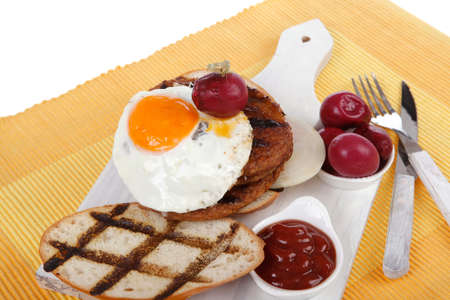 junk food meat big beef hamburger fried eggs on white wood plate with cutlery ketchup sauce and pickels on yellow mat isolated over white backgroundの写真素材