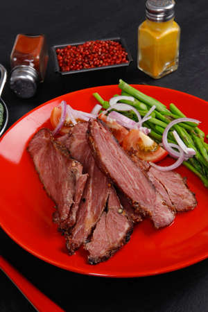 fresh roast beef meat slices on red plate with paprika pink peppercorn hot chili pepper asparagus mustard powder boiled broccoli and cutlery on black wood plateの写真素材