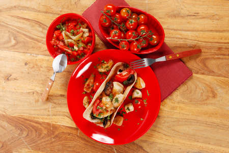 big mexican taco with tomatoes and mushroom, served with soup, fresh vegetables , on red crockery plates over wooden table with cutlery and napkinの写真素材