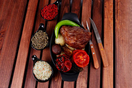 fresh roast bbq beef steak on black pan baked with mushroom potatoes tomatoes green hot chili pepper served with different peppercorn on dark wooden tableの写真素材