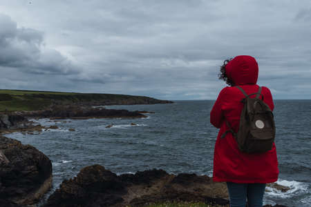 Girl in a red coat with bag looking at the sea in a cloudy dayの写真素材