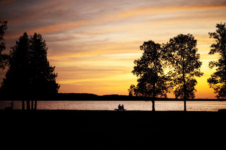 Elderly couple, silhouetted,  sitting on a bench by lake at sunsetの写真素材