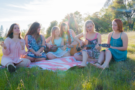 Young Girls Sitting Together in Grassy Field Singing and Playing Musical Instruments With Sunlight Overheadの写真素材
