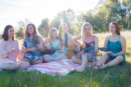 Young Girls Sitting Together in Grassy Field Singing and Playing Musical Instruments With Sunlight Overheadの写真素材