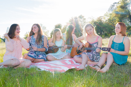 Young Girls Sitting Together in Grassy Field Singing and Playing Musical Instruments With Sunlight Overheadの写真素材