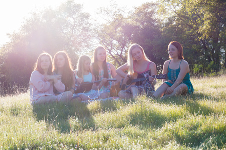 Young Girls Sitting Together in Grassy Field Singing and Playing Musical Instruments With Sunlight Overheadの写真素材