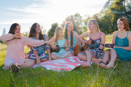 Young Girls Sitting Together in Grassy Field Singing and Playing Musical Instruments With Sunlight Overheadの写真素材