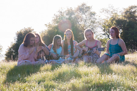 Young Girls Sitting Together in Grassy Field Singing and Playing Musical Instruments With Sunlight Overheadの写真素材