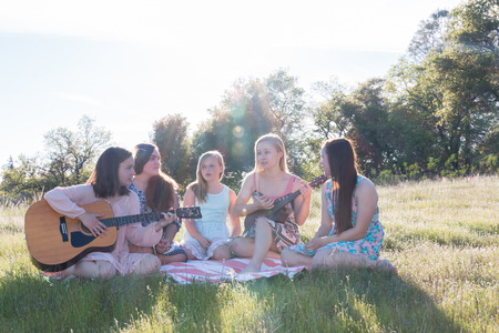 Young Girls Sitting Together in Grassy Field Singing and Playing Musical Instruments With Sunlight Overheadの写真素材