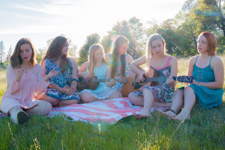 Young Girls Sitting Together in Grassy Field Singing and Playing Musical Instruments With Sunlight Overheadの写真素材