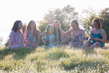 Young Girls Sitting Together in Grassy Field Singing and Playing Musical Instruments With Sunlight Overheadの写真素材