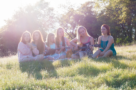 Young Girls Sitting Together in Grassy Field Singing and Playing Musical Instruments With Sunlight Overheadの写真素材
